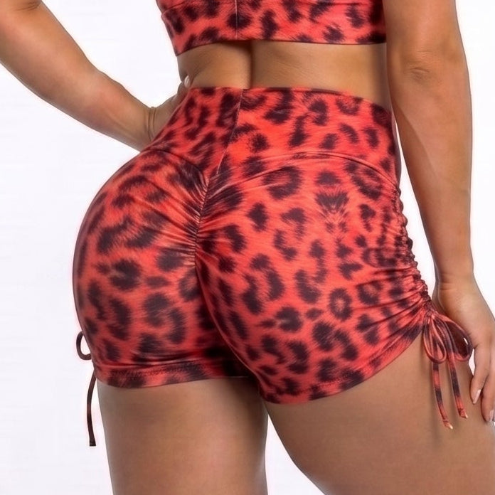 A woman modeling a red and black leopard print activewear set, featuring a sports bra and high-waisted shorts, shown from a rear three-quarter view against a white studio background.